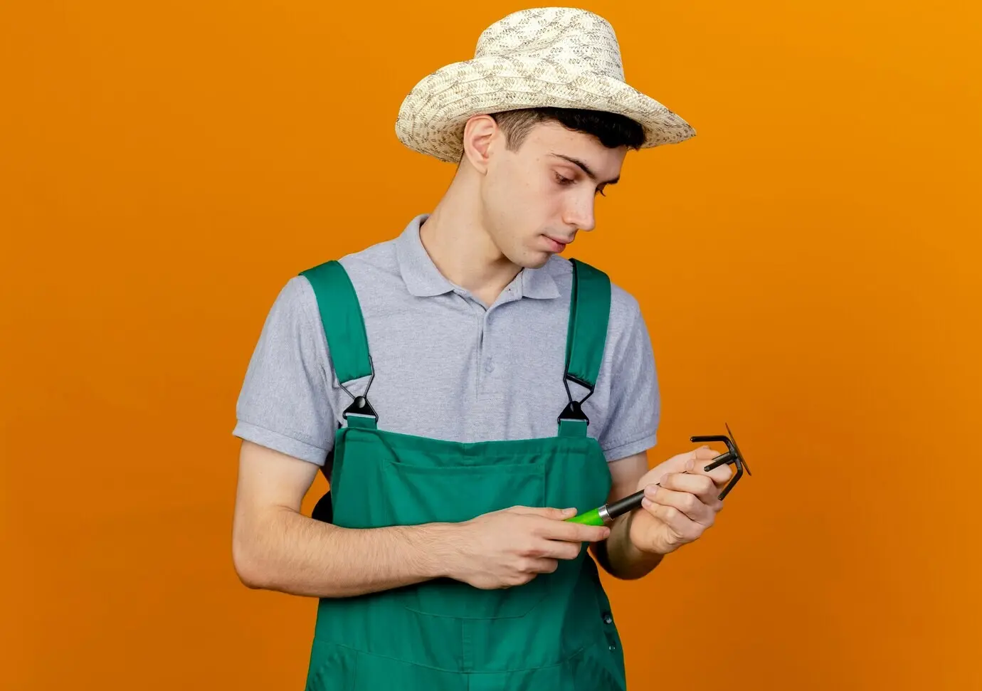 Selbstbewusster junger männlicher Gärtner mit Gartenhut blickt auf eine Hacke-Harke.