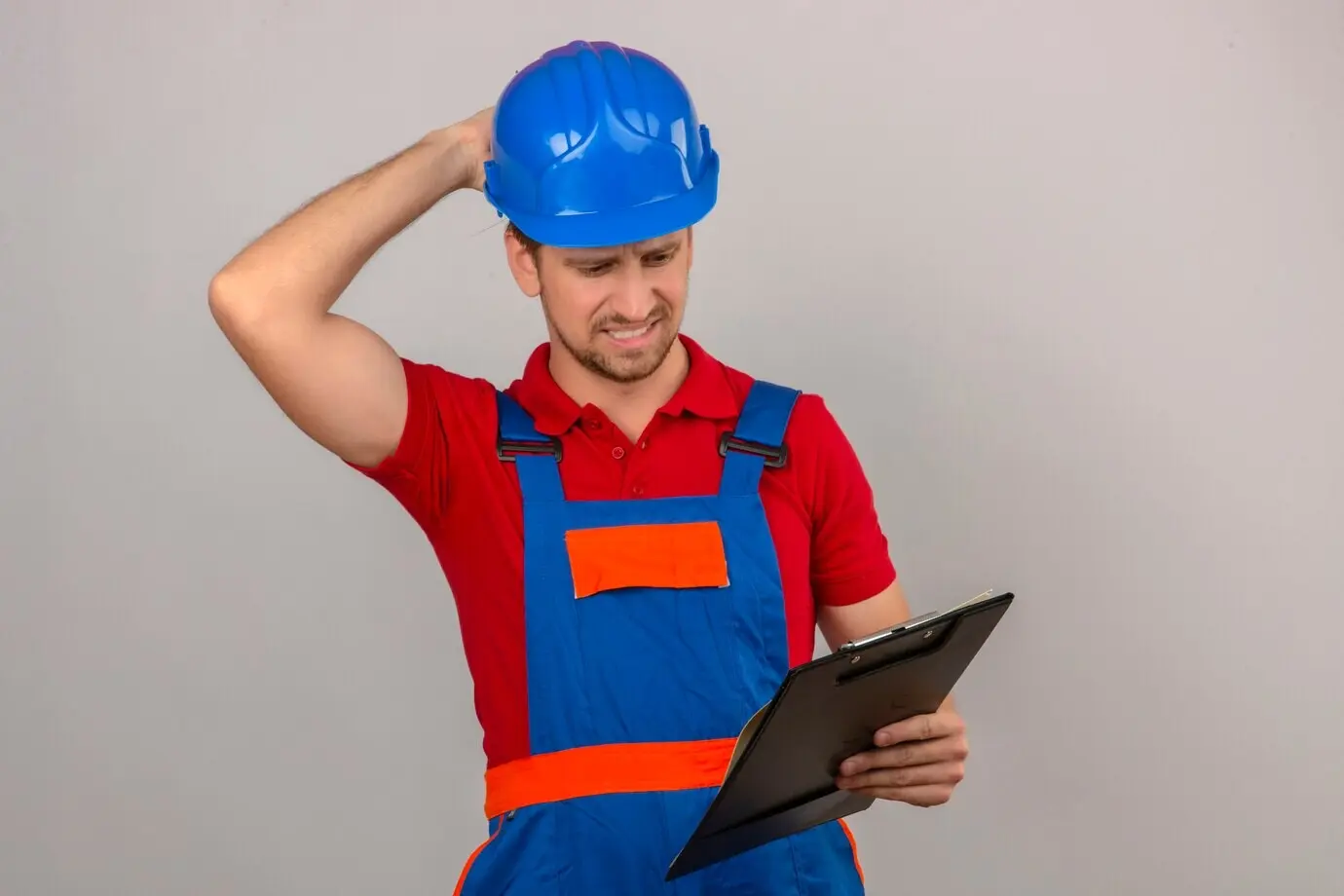 Junger Bauarbeiter in Baukleidung und Schutzhelm, hält ein Klemmbrett, nachdenklich mit der Hand am Kopf, mit verwirrtem, zweifelndem Blick, vor einer isolierten weißen Wand.
